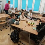 Dzieci siedza przy stole w klasie, leżą na nim talerze z kanapkami. Dzieci jedzą kanapki.