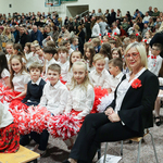 Grupa uczniów ubranych na biało-czerwono siedzi na sali gimnastycznej z nauczycielką. W tle tłum ludzi.
