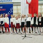 Na sali gimnastycznej stoją uczniowie ubrani w barwy biało-czerwone oraz stroje czarno- białe. W tle flaga polska oraz na grantowym tle napis Koncert dla niepodległej..