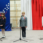 Chłopiec w garniturze oraz dwie dziewczynki stoją przed mikrofonem, w tle barwy biało-czerwone.