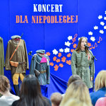 Dziewczyna stoi przed mikrofonem i śpiewa, w tle widać mundury żołnierskie oraz napis Koncert dla niepodległej na granatowym tle.