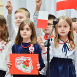 Zdjęcie dzieci ubranych na biało i czerwono. Dziewczynki trzymają flagi oraz rysunek. 