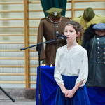 Dziewczynka stoi przed mikrofonem, w tle widać mundury żołnierskie.