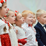 Zdjęcie  śpiewających dzieci ubranych na biało i czerwono. Dziewczynki mają wianki na głowach. 
