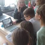 Uczniowie stoją przy biurku nauczyciela i nachylają się nad laptopem.