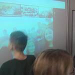 Uczniowie stoją przy biurku nauczyciela i patrzą na tablicę multimedialną