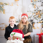 Dziewczynka w mikołajowej czapce i chłopiec trzymają poduszkę w kształcie głowy Mikołaja.  W tle bożonarodzeniowy wystrój
