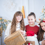 Trzy dziewczynki pozują do zdjęcia, trzymają paczki świateczne. W tle bożonarodzeniowy wystrój.