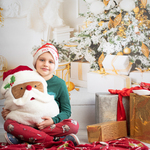 Chłopiec siedzi w mikołajowej czapce z poduszką w kształcie głowy Mikołaja.  W tle bożonarodzeniowy wystrój