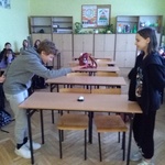 Dwoje uczniów stoi naprzeciwko siebie przy ławce w klasie. Jedną ręką naciskają dzwonek, który stoi na ławce.