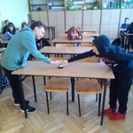 Dwoje uczniów stoi naprzeciw siebie przy ławce w klasie. Jedną ręką naciskają dzwonek, który stoi na ławce..