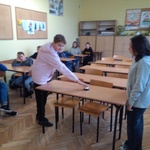 Dwoje uczniów stoi naprzeciw siebie przy ławce w klasie. Jedną ręką naciskają dzwonek, który stoi na ławce.
