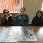 Trzy dziewczyny siedzą przed dużym banknotem.jpg