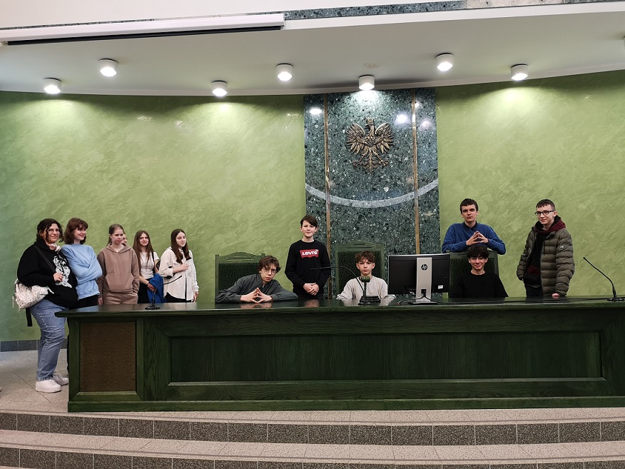 Uczniowie siedzą i stoją przy dużym zielonym biurku sędziowskim w sali sądu..jpg