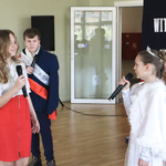 Dziewczynka ubrana na biało mówi do mikrofonu. Na przeciwko niej stoi dwoje uczniów ubranych na galowo. W tle patriotyczny napis.