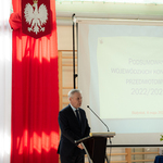 Pan dyrektor przemawia z mównicy, w tle flaga polska oraz ekran z napisami..
