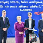 Przedstawiciele władz oświatowych stoją razem z uczniem, który trzyma dyplom i papierową torbę. -