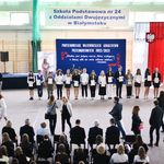 Grupa uczniów stoi z dyplomami na tle napisów na niebieskim płótnie.