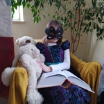 Dziewczynka przebrana w suknię księzniczki oraz z maską na twarzy czyta książkę. W ręku trzyma obok siebie misia..jpg