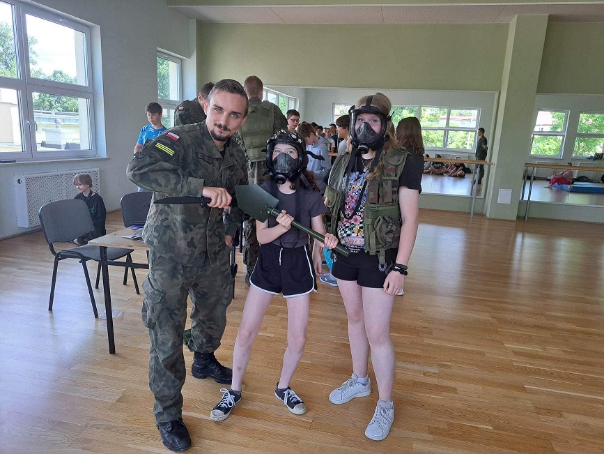 Żołnierz trzyma w ręku karabin. Obok stoją dwie dziewczyny ubrane w maski przeciwgazowe oraz wojskowe kamizelki..jpg