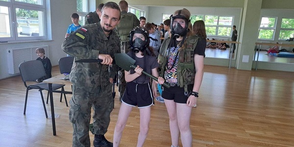 Żołnierz trzyma w ręku karabin. Obok stoją dwie dziewczyny ubrane w maski przeciwgazowe oraz wojskowe kamizelki..jpg