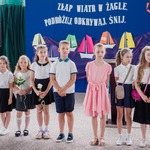 Młodsi uczniowie ubrani na glaowo stoją przed tablicą z napisami. Troje z nich trzyma róże.