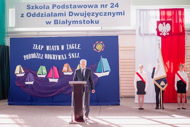 Pan dyrektor przemawia przy mównicy. Obok stoi poczet sztandarowy na tle flagi biało-czerwonej..jpg