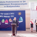 Pan dyrektor przemawia przy mównicy. Obok stoi poczet sztandarowy na tle flagi biało-czerwonej.