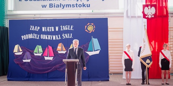 Pan dyrektor przemawia przy mównicy. Obok stoi poczet sztandarowy na tle flagi biało-czerwonej..jpg