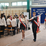 Uczniowie ubrani w stroje biało -czarne wychodzą ze sztandarem za sali gimnastycznej.