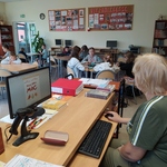 Uczniowie siedzą w ławkach i przy komputerze.
