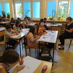 Uczniowie siedzą w ławkach, nauczyciel stoi na środku sali.