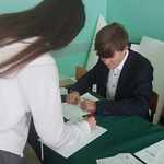 elegancko ubrany uczeń siedzi przy stole. Na przeciwko dziewczyna podpisuje listę