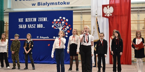 Grupa uczniów ubranych w stroje galowe występuje na uroczystej akademii. W tle flaga Polski oraz dekoracja patriotyczna na niebieskim płótnie.