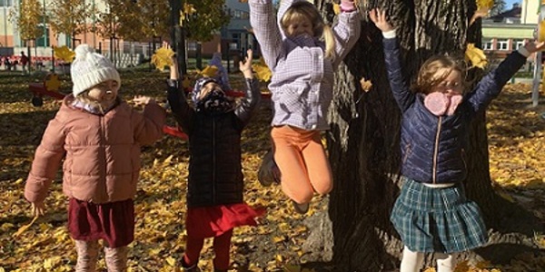 Cztery dziewczynki stoją przy drzewie i podrzucają do góry kolorowe liście.