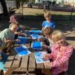 Grupa uczniów siedzi przy ławie na podwórku i maluje na niebiesko kartki..jpg