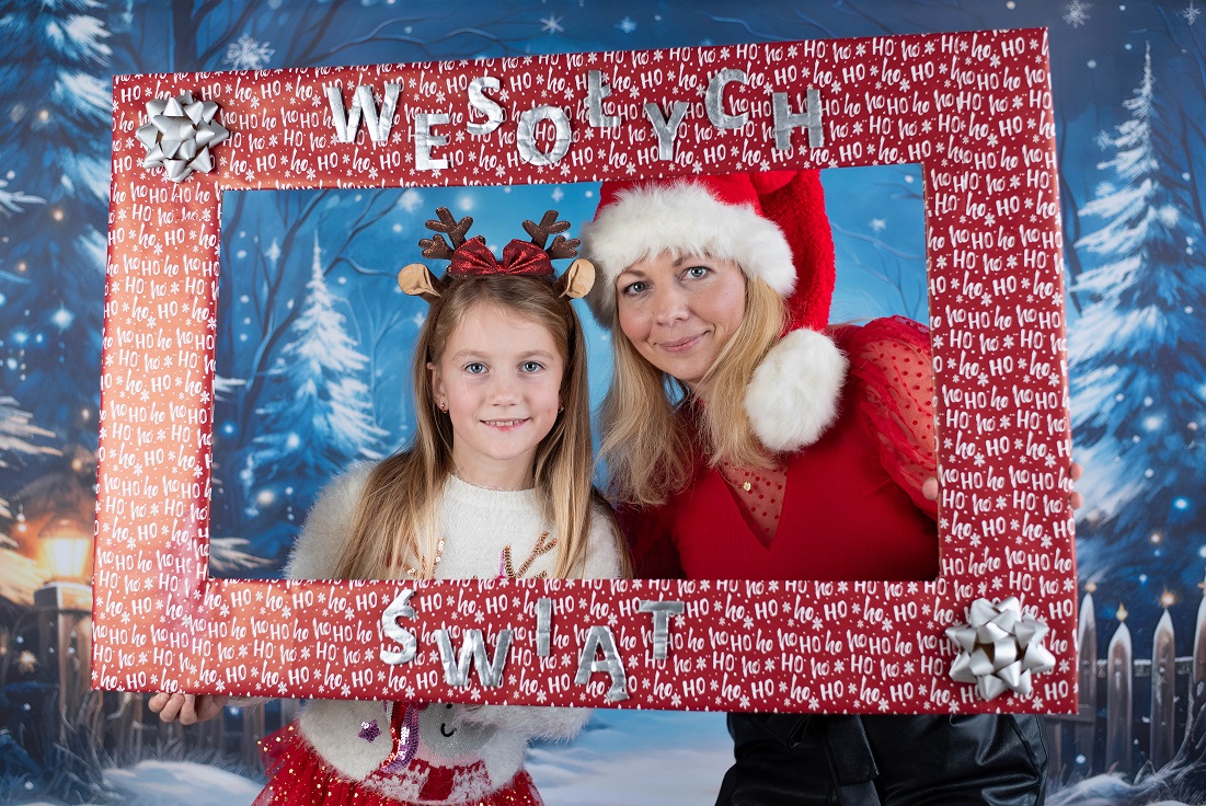 Pani ubrana w strój Mikołaja oraz dziewczynka w opasce z reniferem w świątecznej fotobudce.