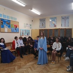 Dziewczyna w błękitnej sukni stoi na tle uczniów siedzących w sali..jpg