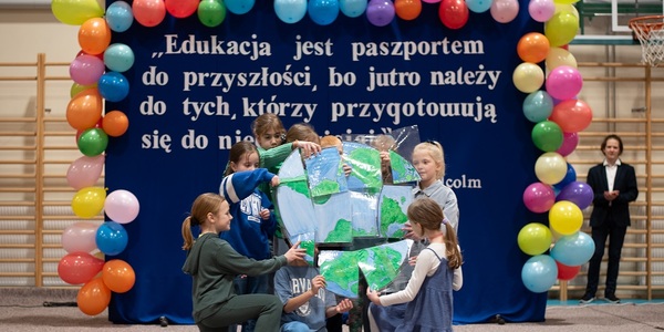 Grupa dzieci trzyma fragmenty rysunku. Za nimi tablica z napisami i kolorowymi balonami..jpg