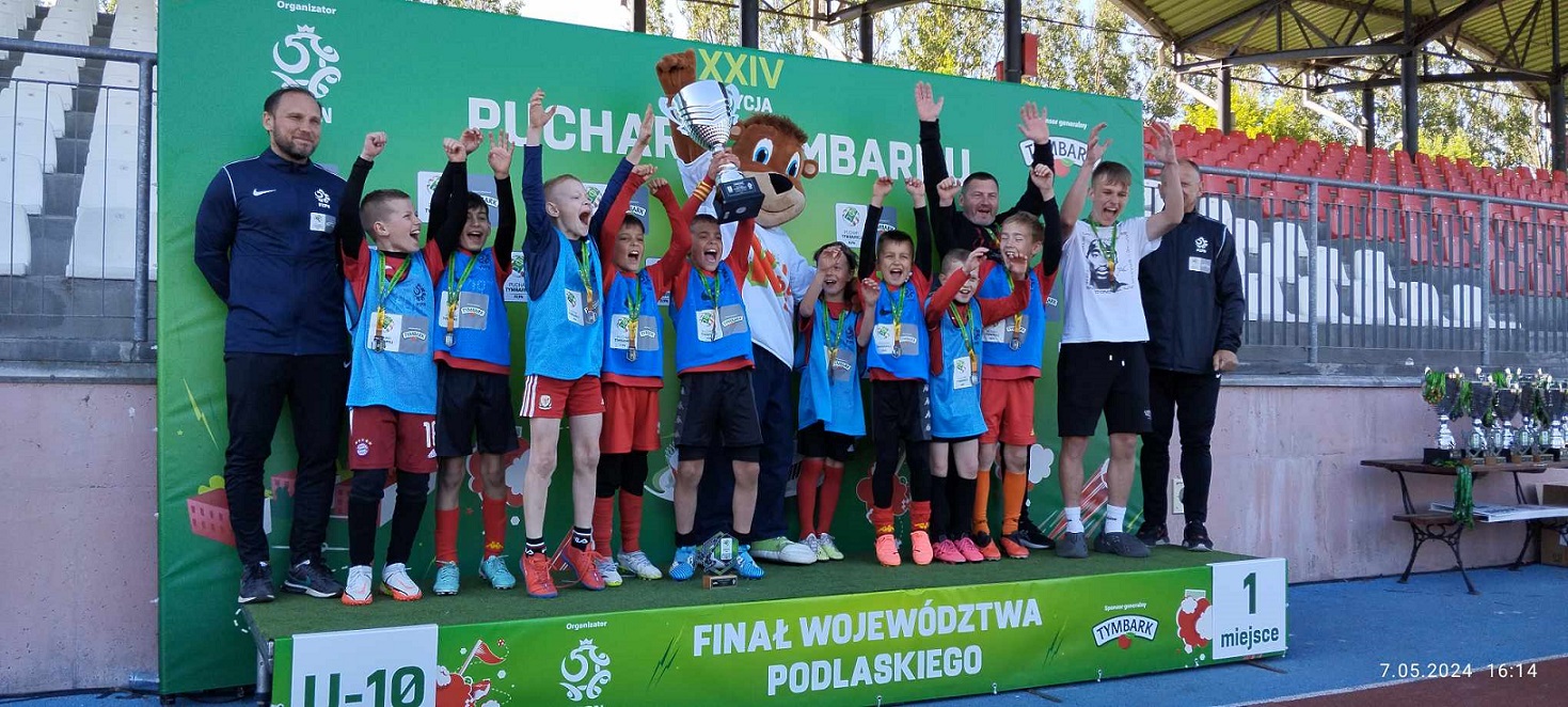 Chłopcy w sportowych strojach stoją na podium i trzymają ręce w górze oraz puchar.jpg