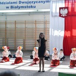 Dziewczynka występuje w przedstawieniu, w tle niebieska tablica z czerwonymi makami oraz flaga polska