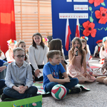 Grupa dzieci występuje w przedstawieniu, w tle niebieska tablica z czerwonymi makami oraz flaga polska