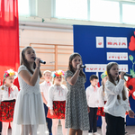 Dziewczynki śpiewają, w tle niebieska tablica z czerwonymi makami oraz flaga polska