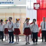 Dzieci ubrane w tradycyjne stroje ludowe stoją na sali, w tle tablica z czerwonymi makami oraz flaga Polski.