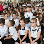 Duża grupa dzieci w strojach galowych siedzi na sali gimnastycznej.