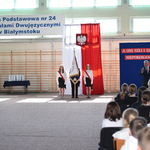 Troje uczniów stoi ze sztandarem, w tle flaga biało-czerwona oraz niebieska tablica z czerwonymi makami.
