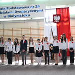 Grupa uczniów w strojach galowych i z czerwonymi makami stoi na tle flagi biało-czerwonej.