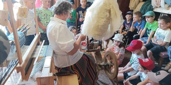 Grupa dzieci siedzi przy pani_ która przedzie wełnę na kołowrotku..jpg