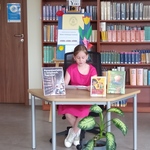 Uczennica czyta książkę przy stoliku z innymi książkami  na tle półek bibliotecznych..jpg