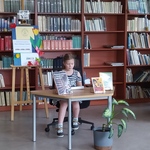Uczennica czyta książkę przy stoliku z innymi książkami  na tle półek bibliotecznych.jpg